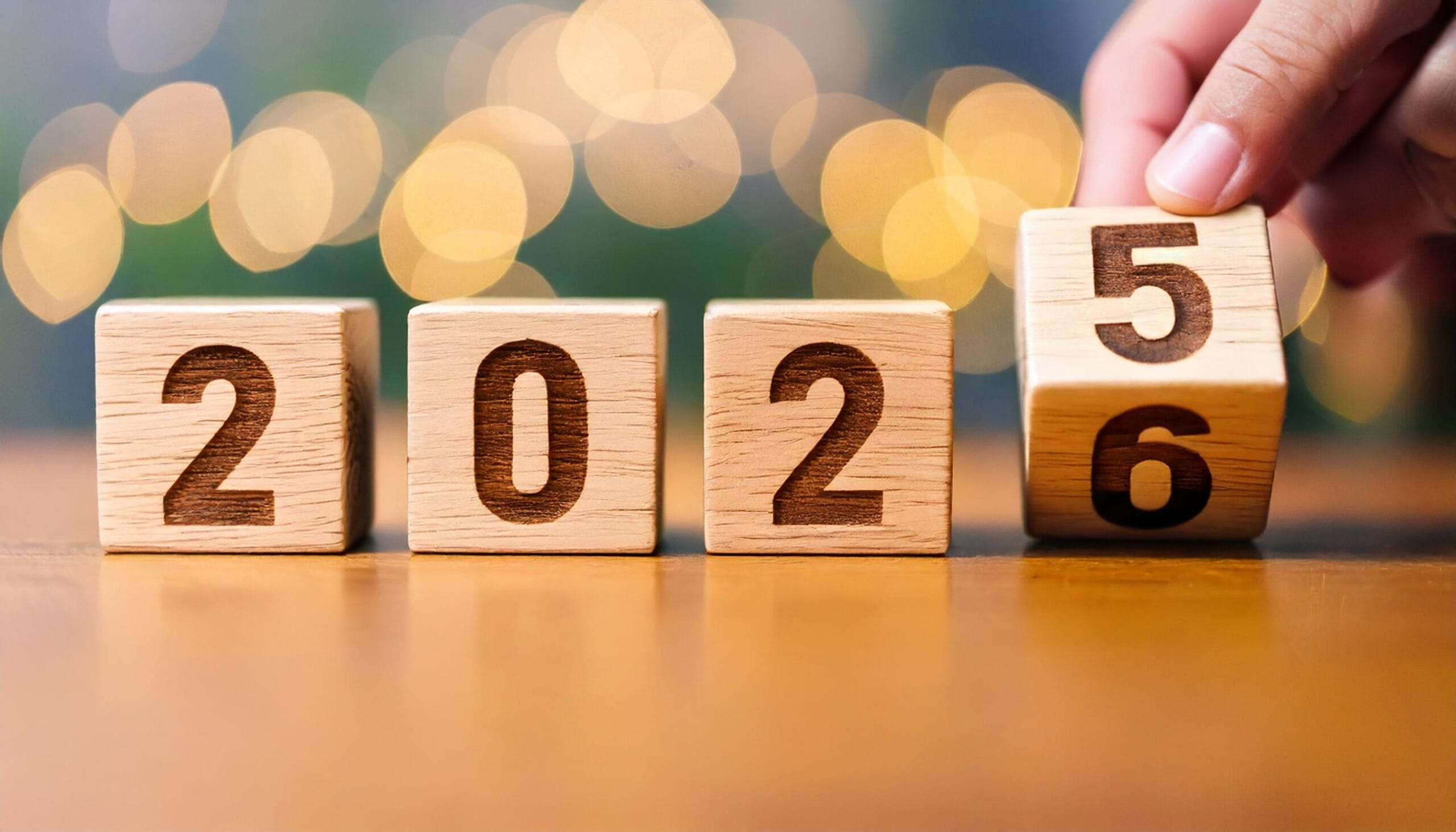 Hand flipping wooden cube from 2025 to 2026 signifying the transition to the new year with bokeh lights in the background