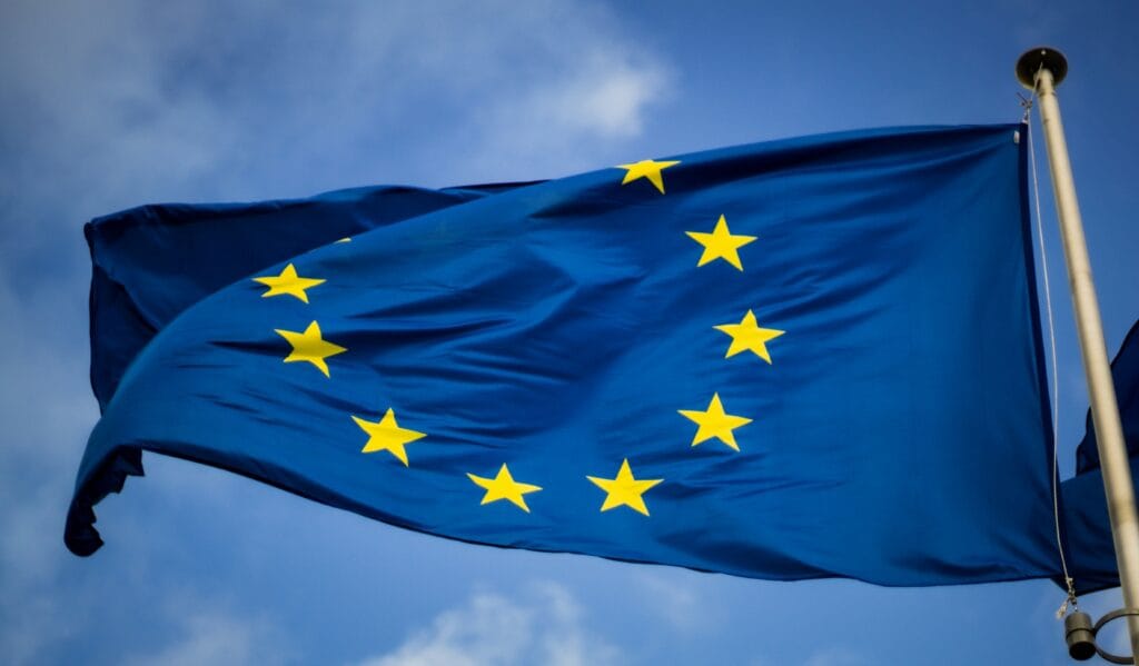 A blue EU flag flies in the wind on a white flagpole, under a blue sky