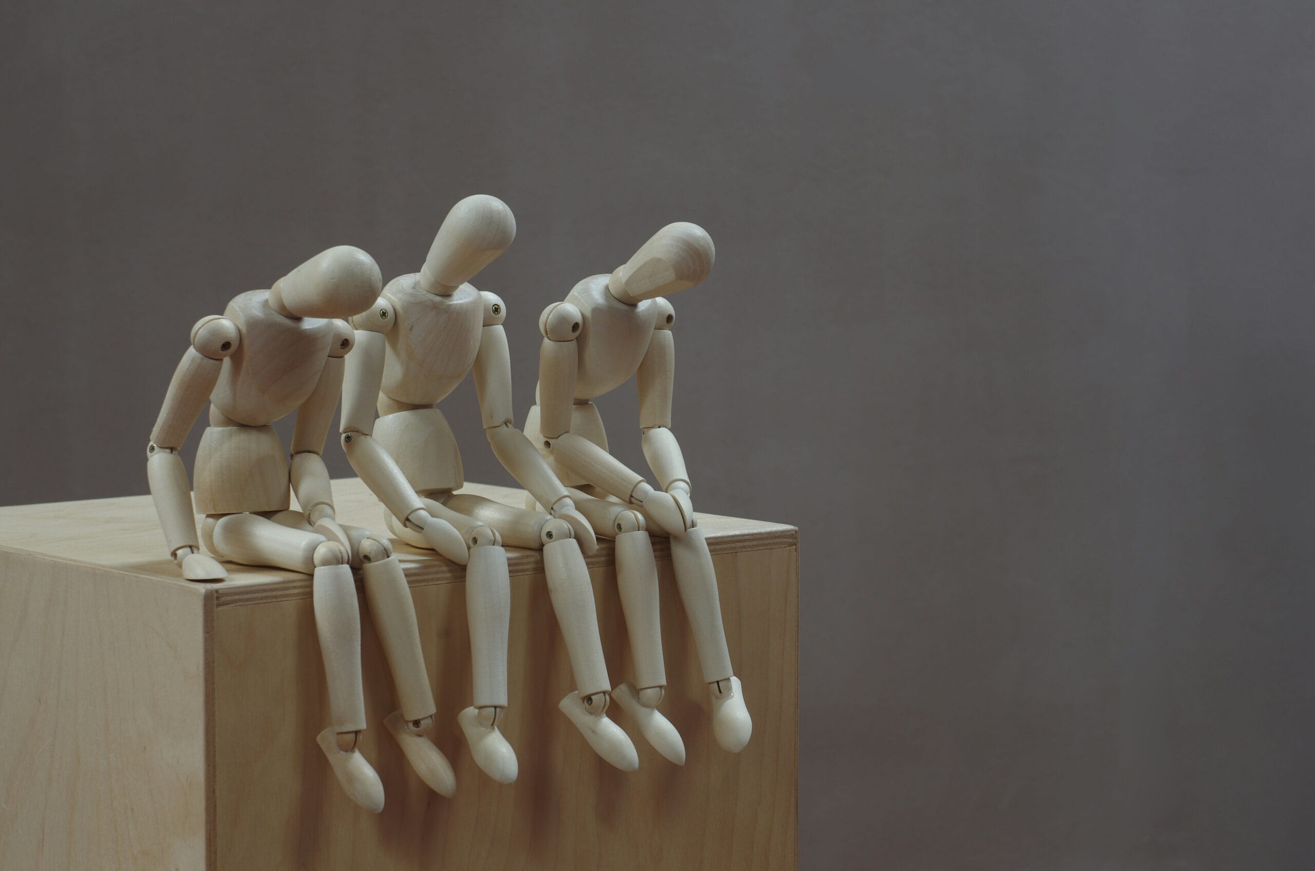 Three art mannequins sat on the edge of a wooden block in front of a beige background